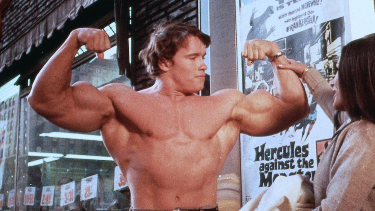 Ein Bodybuilder posiert mit nacktem Oberkörper, während eine Frau ihn am Arm fasst.