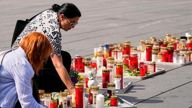 Auf dem Boden stehen einige Kerzen und Blumenkränze, zwei Frauen stellen Kerzen dazu.