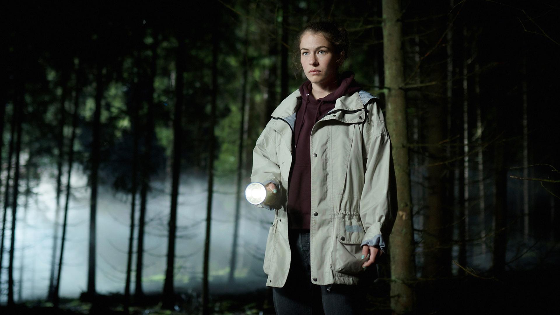 Eine junge Frau mit einer Taschenlampe in einem nächtlichen Wald.