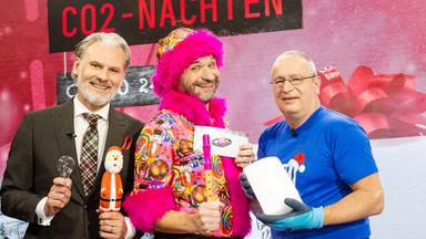 Das Bild zeigt drei Männer, die vor einem festlich dekorierten Hintergrund stehen. An der Wand sind mehrere Weihnachtslichter und ein großes Banner mit der Aufschrift "FROHE CO2-NACHTEN" zu sehen. Der erste Mann, links im Anzug, hält einen Weihnachtsmann in der Hand und eine Glühbirne. Er hat kurzes graues Haar und einen Bart. Der zweite Mann, in der Mitte, trägt ein buntes, auffälliges Outfit mit einem pinken Pelzkragen. Er hält eine Pfeife und ein Schild mit der Aufschrift "Science Busters". Der dritte Mann, rechts, trägt ein blaues T-Shirt und eine Schutzbrille. In seiner Hand hält er einen Block Eis. Alle drei Männer scheinen fröhlich zu sein und lächeln in die Kamera. Der Hintergrund zeigt eine festliche Atmosphäre, die zum Thema Weihnachten passt.