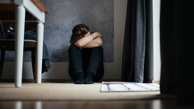 Eine Frau sitzt mit angezogenen Beinen auf dem Boden und versteckt ihr Gesicht.