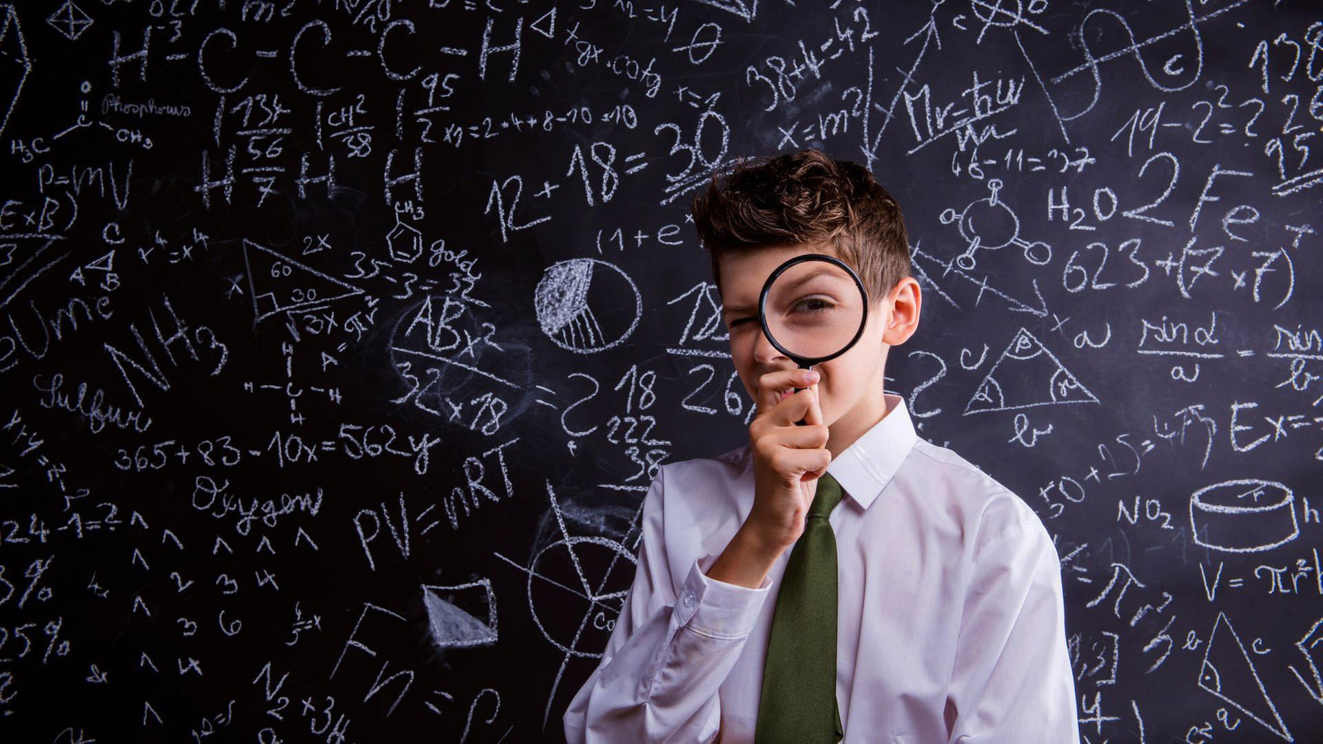 Ein Junge mit Lupe vor dem Gesicht vor einer Tafel mit Formeln und Zahlen.
