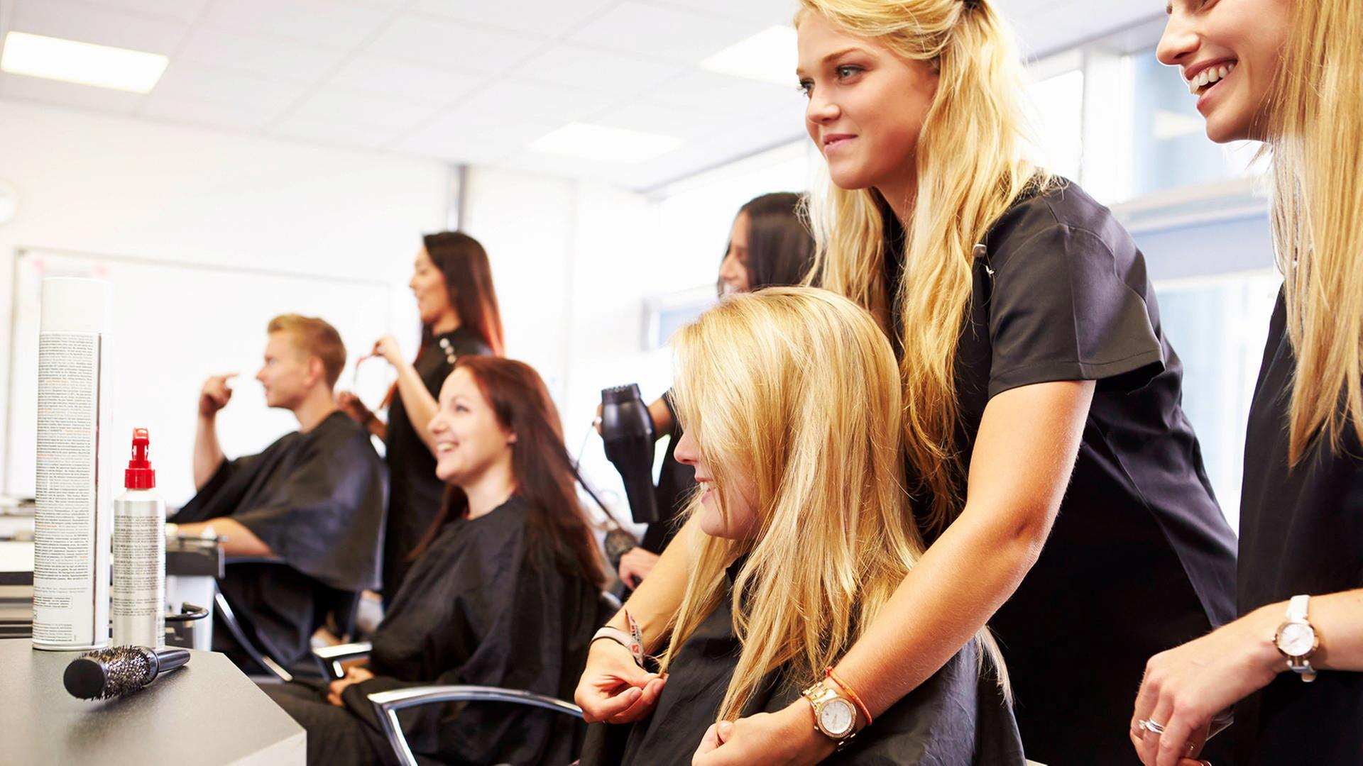 Zwei Frauen und ein Mann sitzen in Friseurstühlen, während hinter ihnen weitere Frauen an ihren Haaren arbeiten.