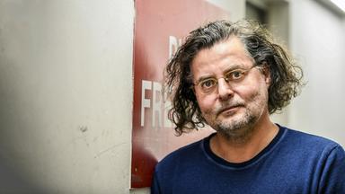 Ein Mann mittleren Alters mit braun-grauem Lockenhaar und Brille steht mit der Schulter an eine Wand gelehnt und blickt mit aufgerissenen Augen in die Kamera.