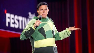 Sebastian23 steht in der Mitte des Bildes. Seine linke Hand streckt er mit der Handinnenfläche nach oben vor sich aus. In der rechten Hand hält er ein Mikrofon. Er trägt einen grünen Pullover und eine Mütze.