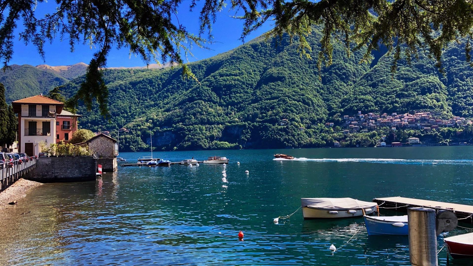 "Seensucht nach Süden - Die oberitalienischen Seen": Blick auf einen smaragdgrünen See, mit Steg im Vordergrund, Häusern auf der linken Seite und bewaldete Berge im Hintergrund.