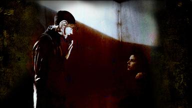  schwarz-weiße historische Filmszene. Sie zeigt zwei Personen vor einer grauen Wand. Eine Person trägt eine Mütze und ein überdimensionales Ohr aus Stoff.