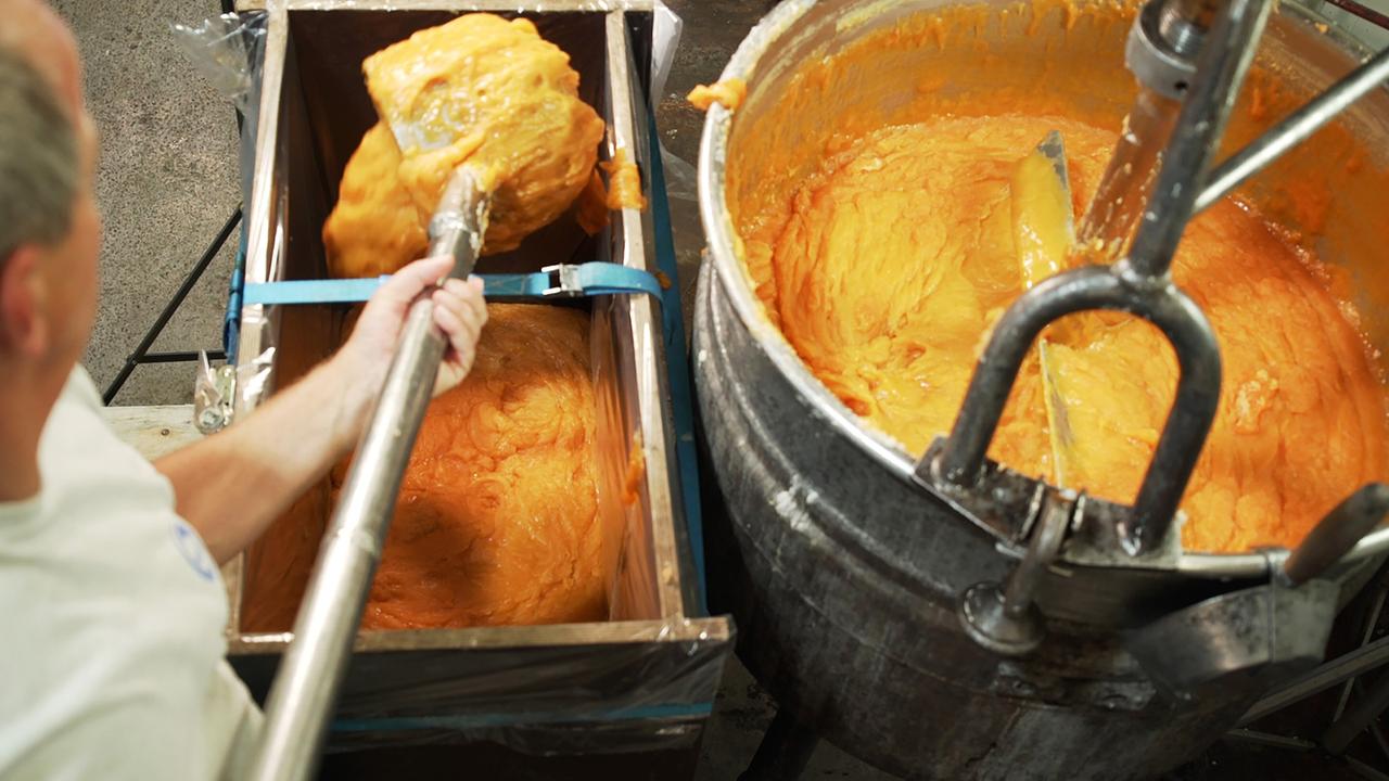 Das Bild zeigt einen Seifensieder in einem Werkstatt-ähnlichen Raum. Im Vordergrund hält eine Person mit einer Hand ein langes Werkzeug, das in einen großen Behälter taucht, in dem eine dickflüssige, orangefarbene Seifenmasse rührt wird. Auf der linken Seite ist ein rechteckiger Behälter sichtbar, der zur Hälfte mit derselben orangefarbenen Masse gefüllt ist und mit einer durchsichtigen Plastikfolie abgedeckt ist. Der Hintergrund ist unscharf, mit Betonboden und Wänden, die auf eine handwerkliche Umgebung hindeuten. Der gesamte Raum wirkt hell erleuchtet.