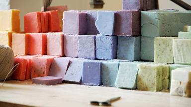 Das Bild zeigt eine Anordnung verschiedener Seifenstücke in unterschiedlichen Farben und Formen. Die Seifen sind in rechteckige Blockform geschnitten und stapeln sich in mehreren Reihen. Die Farben reichen von Gelb, Orange, Pink, Lila bis hin zu Blau und Grün. Einige der Seifenstücke haben eine glatte Oberfläche, während andere kleine Einschlüsse oder Texturen aufweisen, die auf die verwendeten Inhaltsstoffe hinweisen. Im Vordergrund des Bildes liegt ein kleiner, silberner Löffel, und in der Nähe befindet sich eine dünne, natürliche Schnur, die möglicherweise zum Verpacken der Seifen verwendet wird. Der Hintergrund ist unscharf, und es ist nicht erkennbar, was sich dort befindet.