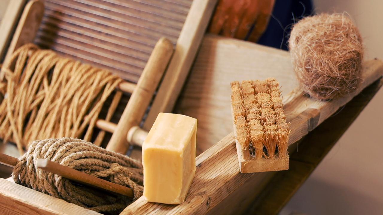 Das Bild zeigt eine Arbeitsfläche, die für die Seifenherstellung genutzt wird. Im Vordergrund liegen ein Stück Seife von gelblicher Farbe sowie ein kleiner Bürstenblock mit vielen feinen Borsten. Daneben befindet sich ein aus kräftigem Material gefertigtes Seil und eine runde, haarnetzartige Struktur aus Naturfaser. Die Arbeitsfläche besteht aus hellem Holz und wirkt rustikal. Im Hintergrund sind weitere Holzmaterialien sichtbar, die möglicherweise Teil des Arbeitsprozesses sind. Die gesamte Anordnung vermittelt einen Eindruck von Handwerk und traditioneller Seifenherstellung.
