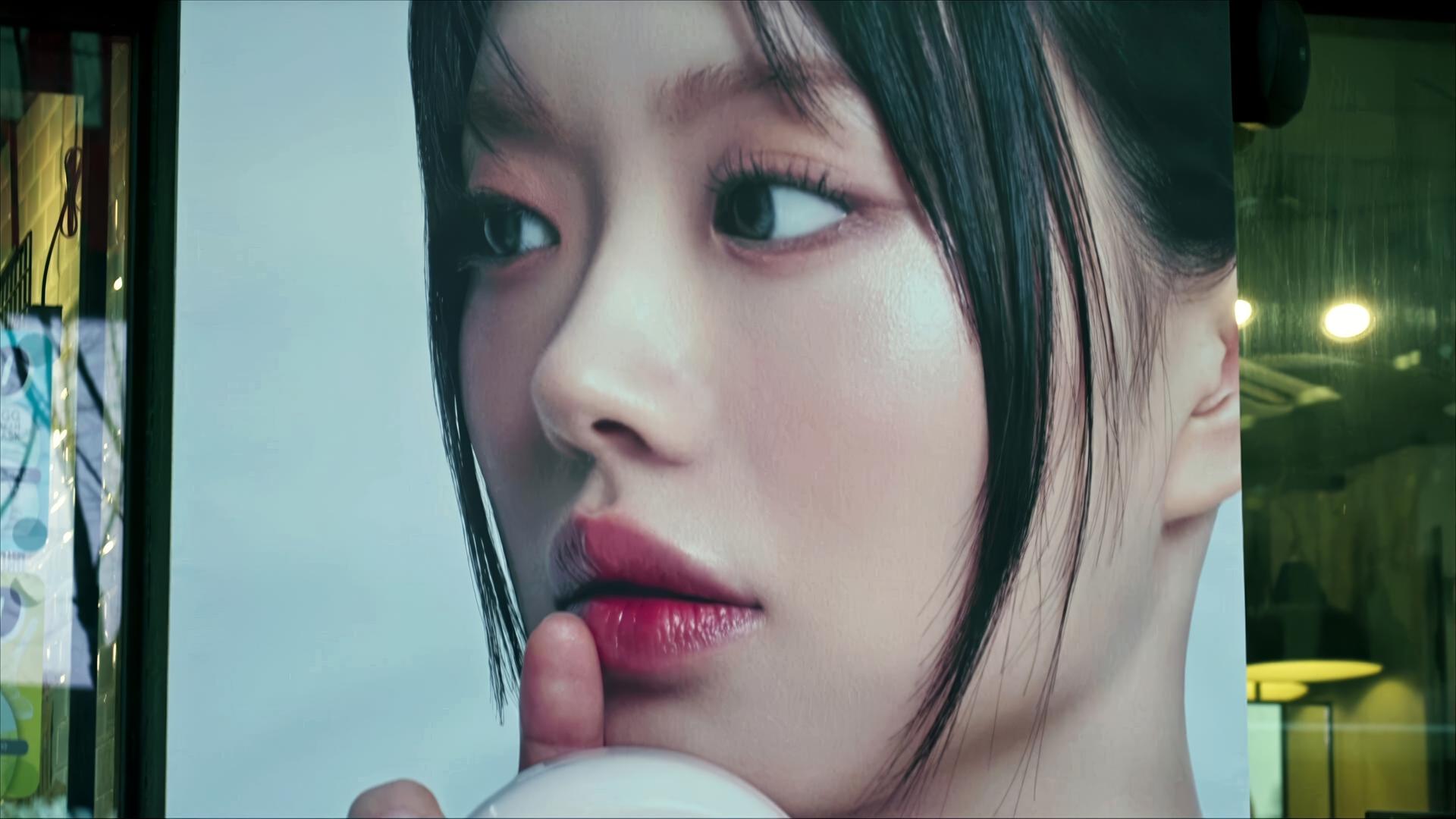 Das Bild zeigt ein großes, nahes Porträt eines Gesichts einer Frau mit glatter, makelloser Haut. Ihr Blick ist direkt nach vorne gerichtet, und ihre Lippen sind leicht geöffnet, als ob sie einen Finger an den Mund legt, was einen nachdenklichen oder geheimnisvollen Ausdruck vermittelt. Die Haare sind dunkel und in einem geraden Schnitt styled, und die Haut hat einen sichtbaren Glanz. Der Hintergrund ist unscharf und in einem hellen Farbton gehalten, was dem Bild eine sanfte und beruhigende Atmosphäre verleiht. Es scheint sich um eine Werbung oder ein Plakat für ein Schönheitsprodukt zu handeln, da das Motiv stark an die K-Beauty-Ästhetik erinnert.