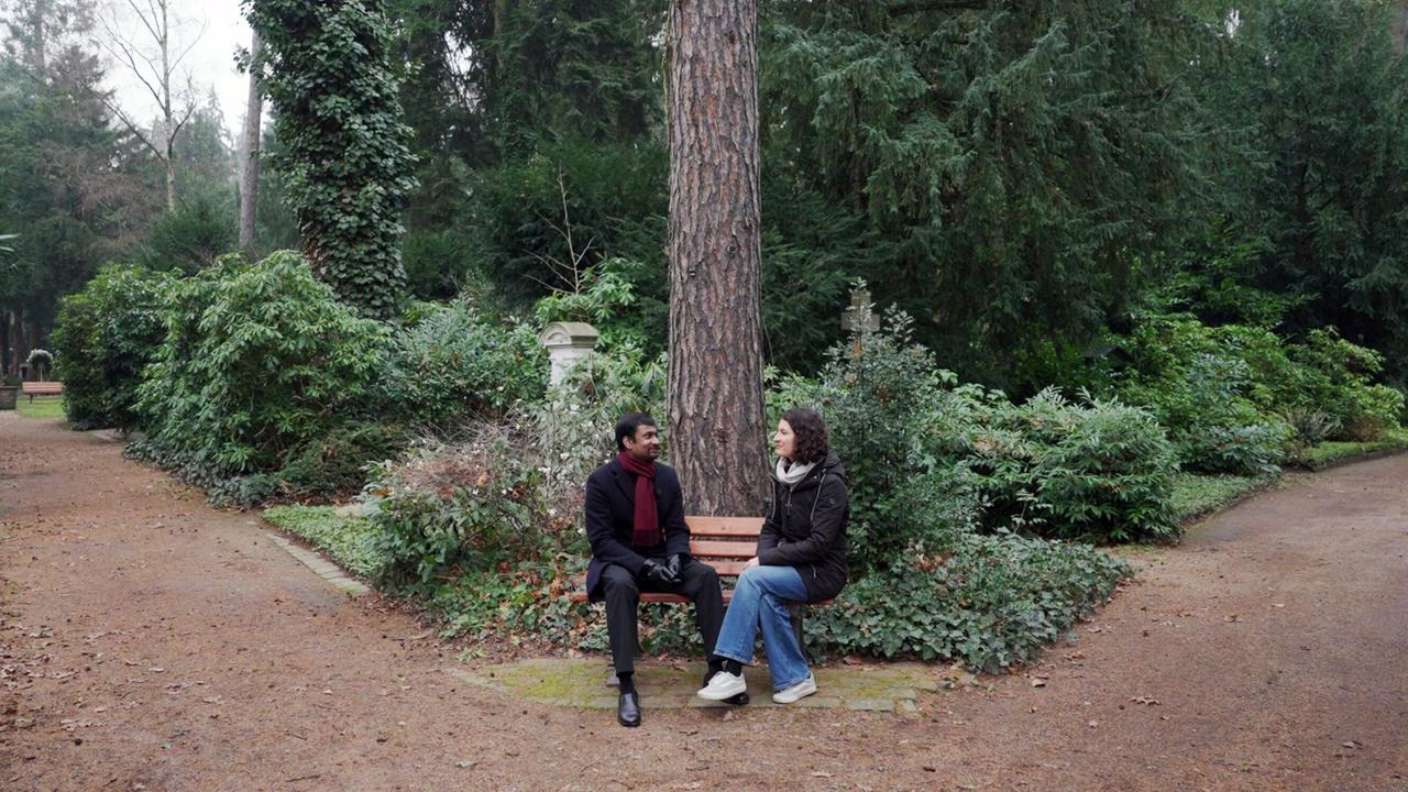 Das Bild zeigt eine ruhige Szene in einem parkähnlichen Bereich, umgeben von üppigem, grünem Bewuchs. Zwei Personen sitzen auf einer Bank in der Mitte des Bildes. Auf der linken Seite ist ein Mann zu sehen, der einen dunklen Mantel trägt und einen roten Schal umgebunden hat. Neben ihm sitzt eine Frau in einem dunklen Mantel und mit einer weißen Jacke. Sie tragen beide legere Kleidung und scheinen ein Gespräch zu führen.  Im Hintergrund sind hohe Bäume und Sträucher zu erkennen, die den Ort eine natürliche Atmosphäre verleihen. Der Boden ist mit Kieselsteinen bedeckt, und es gibt einen leicht sichtbaren Weg, der sich in den Hintergrund zieht. Rechts vom Bild verläuft ebenfalls ein Weg, der durch die Vegetation führt. Die gesamte Szene vermittelt eine ruhige und friedliche Stimmung.