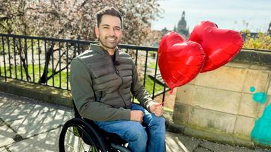 Mann im Rollstuhl vor einer Stadtkulisse mit zwei Herzballons in der Hand