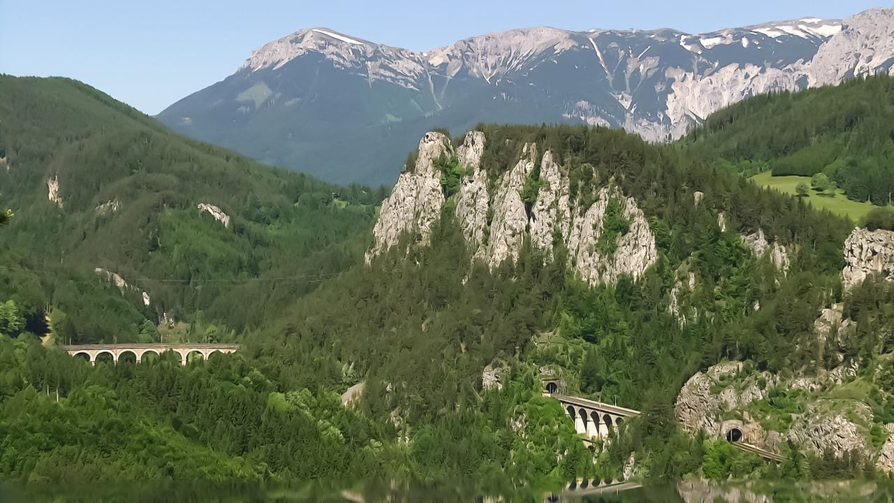 Das Bild zeigt eine beeindruckende Berglandschaft am Semmering, einem Ort im Süden Niederösterreichs. Die Szenerie ist dominiert von grünen Wäldern und steilen Berghängen. Im Vordergrund verläuft ein Viadukt mit mehreren Bögen, das Teil der Semmeringbahn ist, die durch diese malerische Gegend führt. An mehreren Stellen sind Tunnelöffnungen zu sehen. Im Hintergrund erhebt sich ein markanter Berg, der von Wäldern und Wiesen umgeben ist. Der Himmel ist klar und strahlend blau, wodurch die natürliche Schönheit der Landschaft besonders zur Geltung kommt. Zusammenfassend vermittelt das Bild ein Gefühl von Ruhe und idyllichem Naturraum, das für die Region typisch ist.