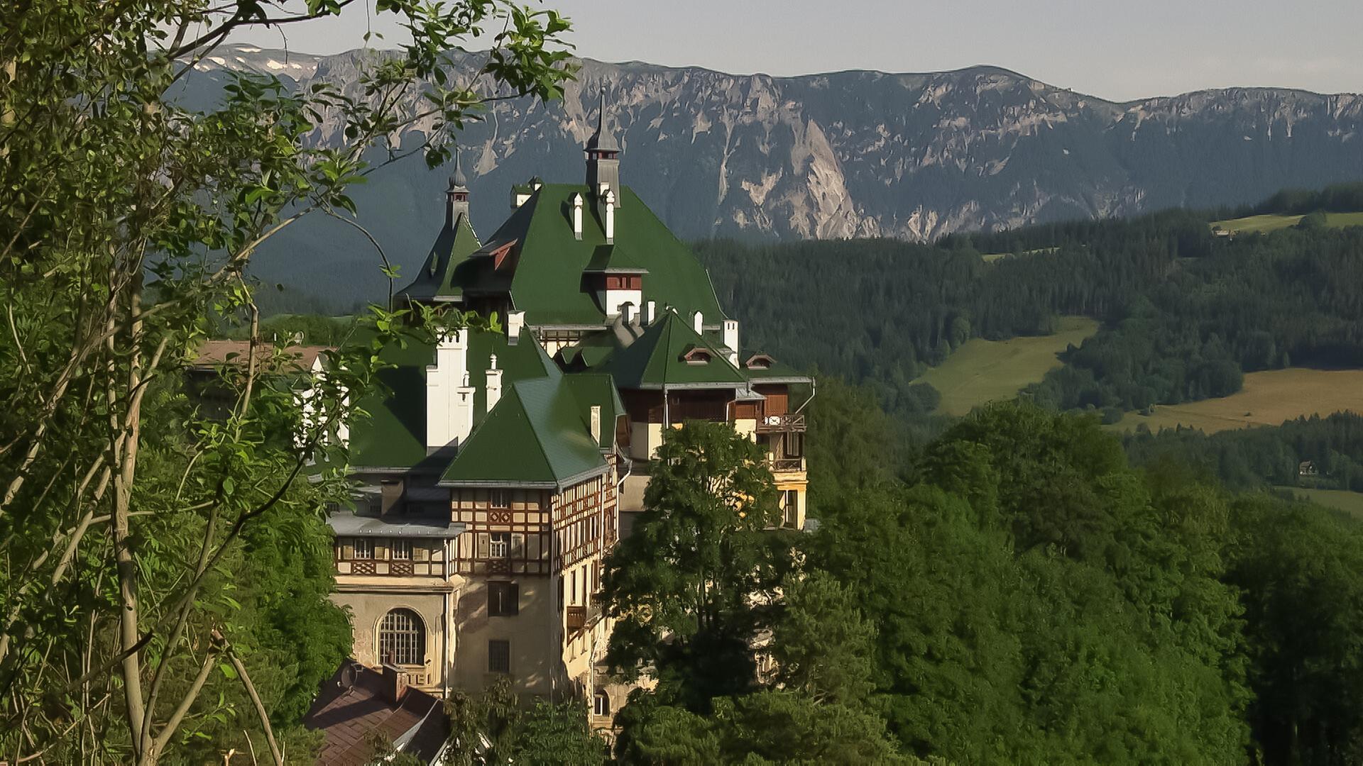 Das Bild zeigt eine hochgelegene Architektur im Semmering, Österreich. Das Gebäude hat mehrere spitz zulaufende Dächer in verschiedenen Grüntönen und ist aus einer Kombination von Stein- und Holzstrukturen konstruiert. Es ist umgeben von dichtem Grün, darunter Bäume, die den Blick auf das Gebäude teilweise verdecken. Im Hintergrund erstrecken sich majestätische Berge mit bewaldeten Hängen und einigen schneebedeckten Spitzen. Der Himmel ist klar und hell, was auf ein sonniges Wetter hinweist. Die Landschaft vermittelt einen Eindruck von natürlicher Schönheit und Ruhe, typisch für das alpine Gebiet.