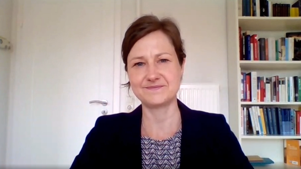 Eine Frau mit brauem Haar und Blazer sitzt in einem Raum vor einem Bücherregal.