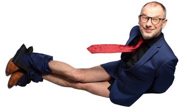 Das Bild zeigt einen Mann, der in einer unkonventionellen Pose auf einer weißen Hintergrundfläche steht. Er trägt einen blauen Anzug mit einem schwarzen Hemd und einer roten Krawatte. Seine Unterhose ist sichtbar, da seine Hose bis zu den Knien heruntergezogen ist. Der Mann hat kurze Haare, eine Brille und ein breites Lächeln auf dem Gesicht, was ihm einen freundlichen Ausdruck verleiht. Seine braunen Schuhe sind gut sichtbar. Die Komposition und der Kontrast zwischen dem Anzug und der weißen Hintergrundfläche betonen den humorvollen Charakter der Pose.