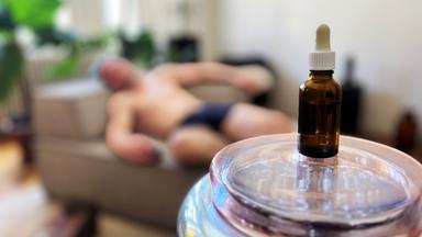 Das Bild zeigt eine nachgestellte Szene, die eine sogenannte Chemsex-Party darstellt. Im Vordergrund des Bildes befindet sich eine braune Glasflasche mit einem Tropfer auf einem gläsernen Tisch. Die Flasche hat einen weißen Deckel. Im Hintergrund ist eine Person zu sehen, die mit freiem Oberkörper auf einem Sofa liegt. Die Person trägt eine kurze Hose und ist offenbar in einem entspannten oder bewusstlosen Zustand. Der Raum hat eine helle, freundliche Atmosphäre mit einer Pflanze im Hintergrund. Der Boden ist aus Holz, und es gibt eine Kiste oder einen kleinen Tisch, der leicht unscharf im Bild zu erkennen ist. Das gesamte Bild vermittelt einen Eindruck von Intimität und der Thematik der Verwendung von Substanzen in einem sexuellen Kontext.