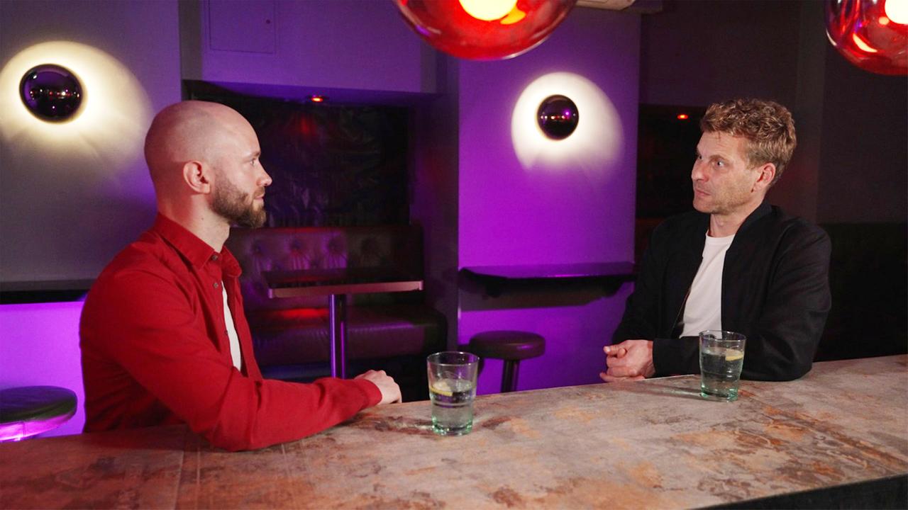 Das Bild zeigt zwei Männer, die an einem Tisch in einer Bar oder einem ähnlichen Lokal sitzen. Der Tisch ist aus Holz und hat eine raue, unebene Oberfläche. Auf dem Tisch stehen zwei Gläser: eines mit klarem Getränk und einer Zitronenscheibe, das andere Glas ähnelt dem ersten und steht in der Nähe einer der Personen.  Der Mann auf der linken Seite hat einen rasierten Kopf, einen kurzen Bart und trägt ein rotes Hemd. Er hat eine nachdenkliche Ausdrucksweise, während er seinen Gesprächspartner ansieht. Der Mann auf der rechten Seite hat lockige, blond-braune Haare und trägt ein dunkles Oberteil. Sein Gesichtsausdruck wirkt angespannt, als wäre er tief in das Gespräch vertieft.  Im Hintergrund sind schwaches, farbiges Licht und eine Couch zu sehen, die vermutlich für das Sitzen von Gästen gedacht ist. Die Umgebung ist düster, mit Lila- und Rottönen, die von verschiedenen Lichtquellen ausgehen. Die Atmosphäre könnte als intim und etwas angespannt beschrieben werden.