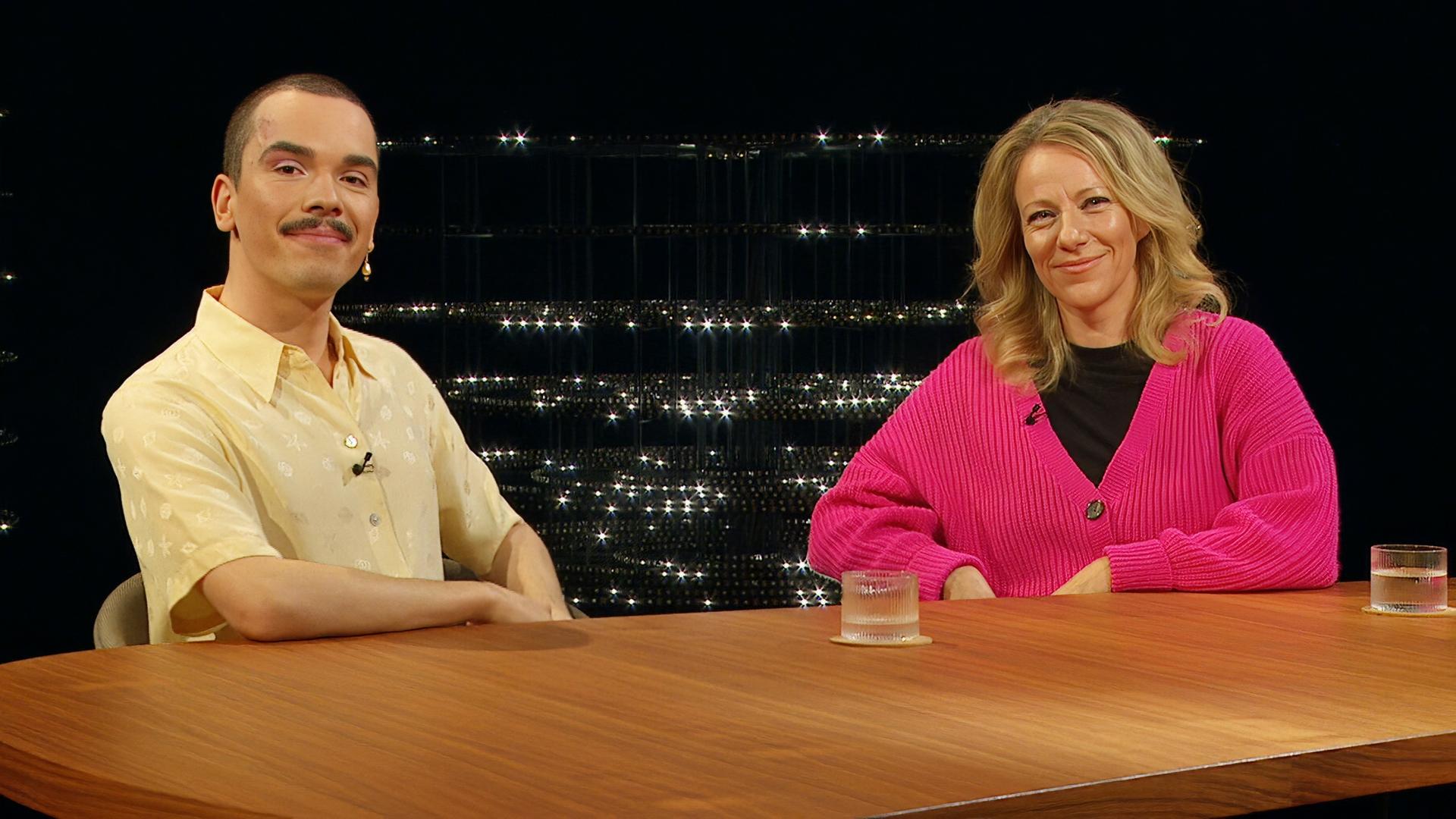 Zwei Personen sitzen an einem Tisch mit Mikrofonen, vor einem dunklen, glitzernden Hintergrund. Eine Person trägt ein gelbes Hemd, die andere einen pinken Pullover.