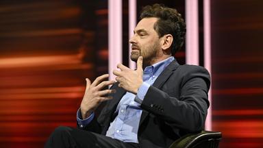 Foto eines Mannes, der in einem Fernsehstudio in einem Sessel sitzt und gestikulierend spricht.