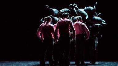 "Gauthier Dance - SWAN LAKES": Mehrere männliche Tänzer in roten Hemden und dunklen Hosen stehen mit dem Rücken zur Kamera. Die Schultern haben alle hochgezogen und die Arme leicht angewinkelt. Über ihren Köpfen aufgetürmt sind schwarze Luftballons in der Form von Schwänen.