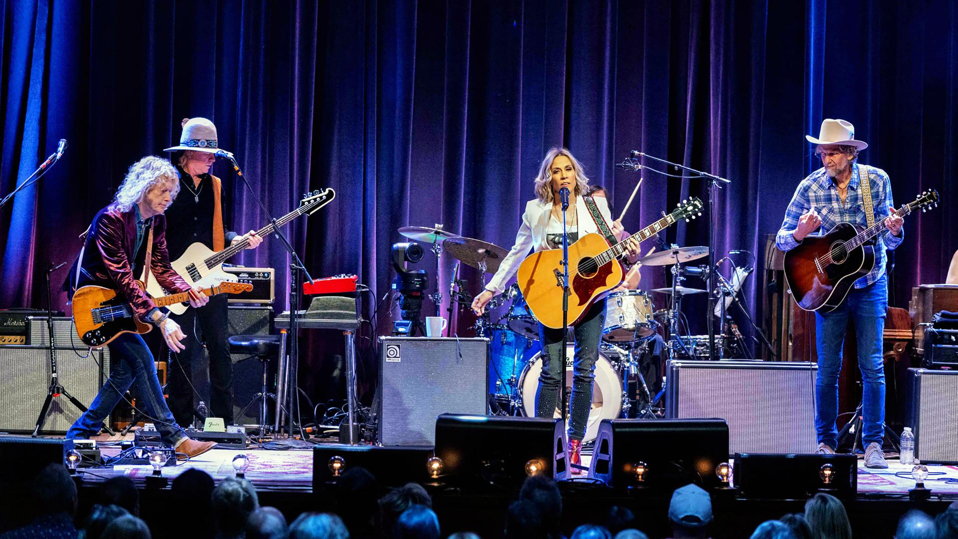 Sheryl Crow steht mit Gitarre und ihrer Band auf der Bühne.