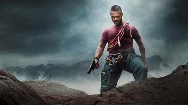 aul Vilar (Philippe Bas) steht auf einem Felsen und schaut in die Tiefe. Er hält eine Pistole in der Hand.