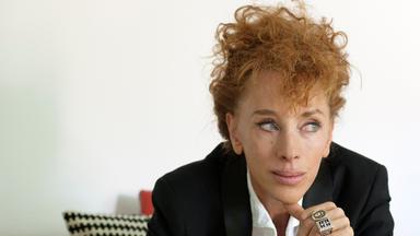 Eine Frau mit wuscheligen roten Haaren hat sich nachdenklich einen Daumen ans Kinn gelegt.