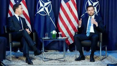 Zwei Männer in Anzügen sitzen nebeneinander auf Stühlen, die vor je zwei Nato- und US-Flaggen aufgestellt wurden, und unterhalten sich.