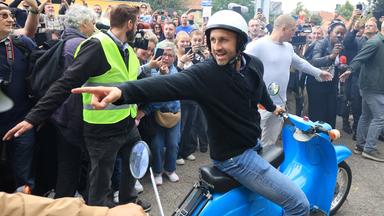 Ein Mann sitzt auf einem blauen Moped und zeigt nach links. Hiter ihm halten zwei Ordner die Menschenmenge davon ab, weiter vor zu treten. 