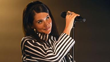Eine junge Frau mit dunklen Haaren und schwarz-weiß gestreiftem Oberteil steht vor einem Mikrofon und lächelt.