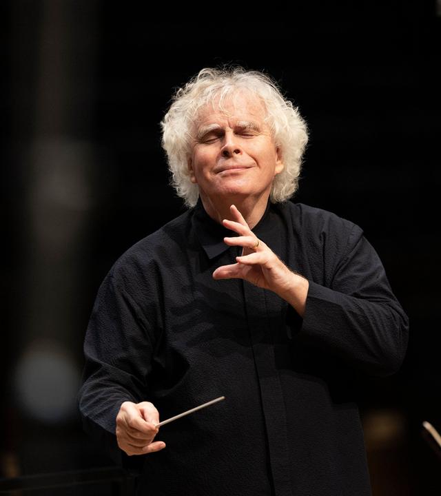 Ein schwarzgekleideter älterer Herr mit Dirigentenstab.