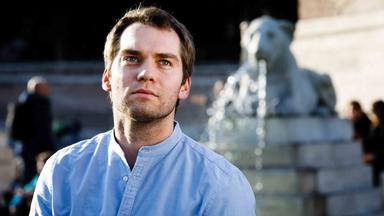 Ein Mann in hellblauem Hemd steht vor einem Brunnen und schaut nach oben über die Kamera hinweg.