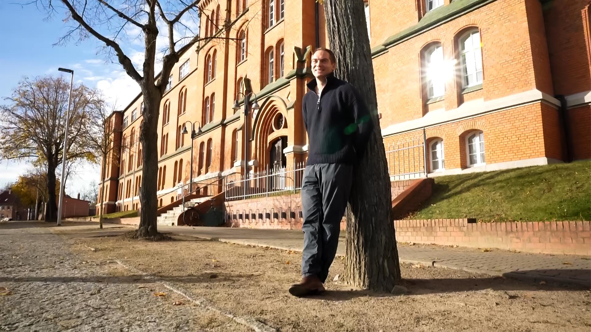 Ein Mann lehnt an einem Baum vor einem alten Backsteingebäude.