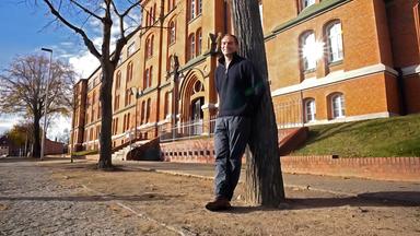 Ein Mann lehnt an einem Baum vor einem alten Backsteingebäude.