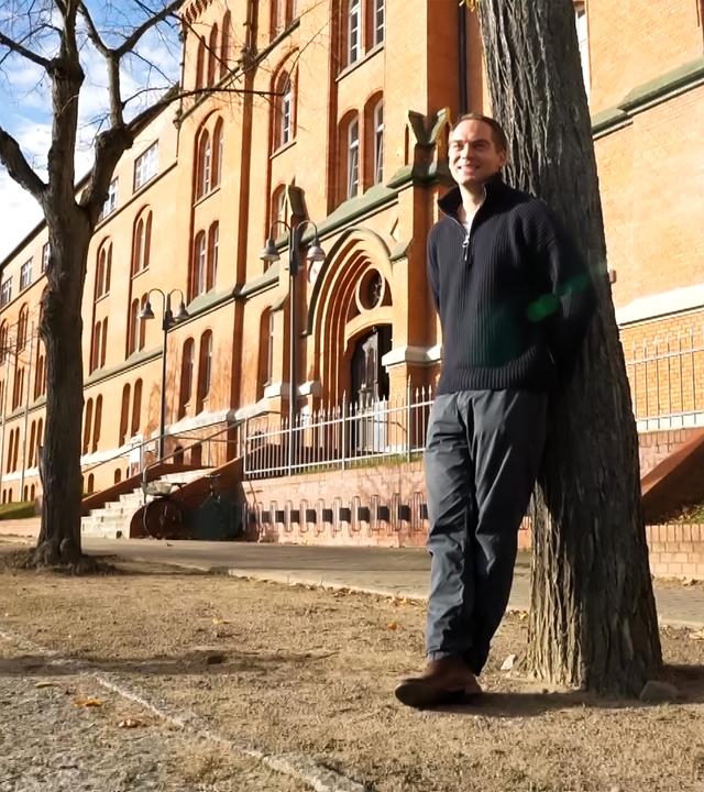 Ein Mann lehnt an einem Baum vor einem alten Backsteingebäude.