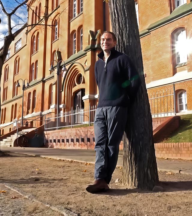 Ein Mann lehnt an einem Baum vor einem alten Backsteingebäude.
