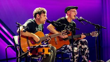 Simon (links) und Jan (rechts) sind auf der Bühne zu sehen. Sie sitzen auf Stühlen in der Mitte des Bildes. Der Hintergrund ist lila beleuchtet.  