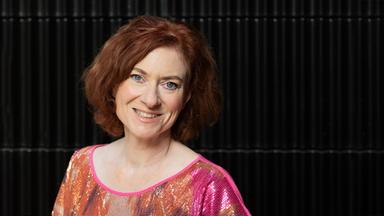 Eine Frau mit schulterlagen braunen wuscheligen Haaren und graublauen Augen trägt ein glitzerndes Pailletten-Shirt in Pinktönen.