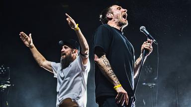 Links im Bild steht ein Mann, mit dunklem Bart und schwarzer Kappe auf dem Kopf. Er trägt ein graues T-Shirt und hebt seine beiden Arme in die Luft. Sein Blick geht nach links. Rechts daneben steht ein weiterer Mann, dessen Blick in die andere Richtung geht und in ein Mikrofon singt. Er trägt ein schwarzes T-Shirt und auf seinem Arm sind Tattoos zu erkennen. Beide stehen auf einer Bühne.