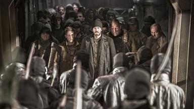 Curtis (Chris Evans) und Edgar (Jamie Bell) stehen in abgerissener Kleidung entschlossen einer Gruppe von Gegnern mit Sturmhauben und Beilen gegenüber, während sich hinter ihnen weitere verwahrlost wirkende Männer in einem dunklen Zugabteil aufreihen.