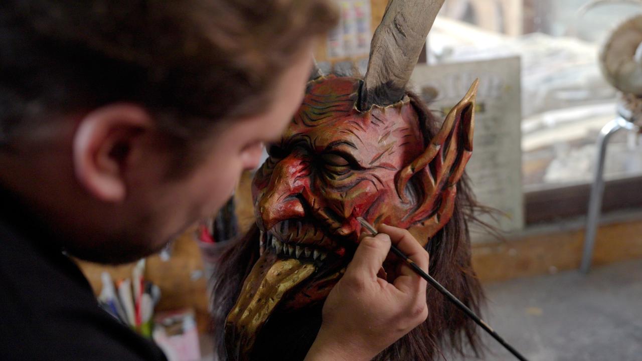 Das Bild zeigt einen Künstler, der mit einem Pinsel an einer fast fertigen Krampusmaske arbeitet. Die Maske ist auffällig gestaltet, mit einer roten Grundfarbe, einem grimmigen Gesichtsausdruck, scharfen Zähnen und langen, nach vorne ragenden Hörnern. Einige Strähnen eines dunklen, haarähnlichen Materials sind an der Maske befestigt.   Der Künstler ist teilweise im Bild zu sehen, sein Gesicht ist jedoch nicht vollständig sichtbar. Er hält den Pinsel mit einer Hand, während die andere Hand die Details der Maske verfeinert. Im Hintergrund sind verschwommene Umrisse von Werkstattmaterialien und eine andere Maske zu erkennen, die darauf hindeuten, dass sich die Szene in einem kreativen Arbeitsraum abspielt.   Die Atmosphäre vermittelt ein Gefühl der Konzentration und handwerklichen Kunstfertigkeit, passend zum Thema des Films, der sich mit traditionellen Weihnachtsbräuchen in Kärnten beschäftigt.