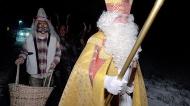 Das Bild zeigt eine nächtliche Szenerie mit mehreren Figuren, die an einem Nikolo-Umzug teilnehmen. Im Vordergrund steht eine Person in einem prächtigen, goldenen Gewand, die eine lange weiße Bartperücke trägt und einen Bischofsstab hält. Diese Figur repräsentiert den Nikolaus, erkennbar an der Mitra, die auf ihrem Kopf sitzt.  Hinter dem Nikolaus befindet sich eine zweite Person, die eine Krampusmaske trägt. Diese Person ist in einen karierten Mantel gekleidet und hält einen Korb in der Hand. Der Krampus, traditionell als Begleiter des Nikolaus, hat eine bedrohliche Maske mit Hörnern und einem furchterregenden Gesichtsausdruck.  Im Hintergrund sind weitere dunkle Figuren zu erkennen, die vermutlich ebenfalls als Krampusse verkleidet sind. Die Umgebung ist dämmerig und der Boden wirkt schneebedeckt. Einige Lichter sind im Hintergrund sichtbar, möglicherweise von Fahrzeugen.  Das Bild vermittelt eine festliche, aber auch mystische Stimmung, geprägt von den Traditionen des Weihnachtsfestes in Kärnten.