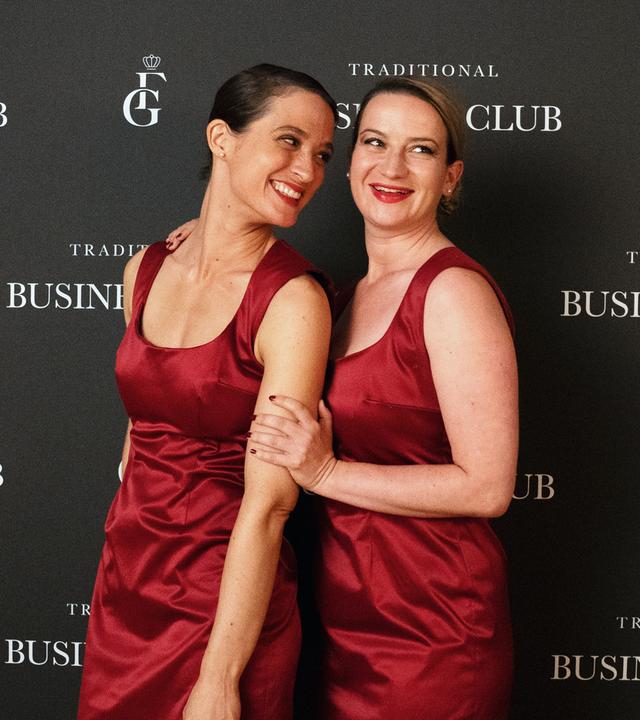 Kim (Friederike Becht) und Maja (Nina Gummich), in roten Cocktailkleidern, stehen lachend vor einer Fotowand eines Events.
