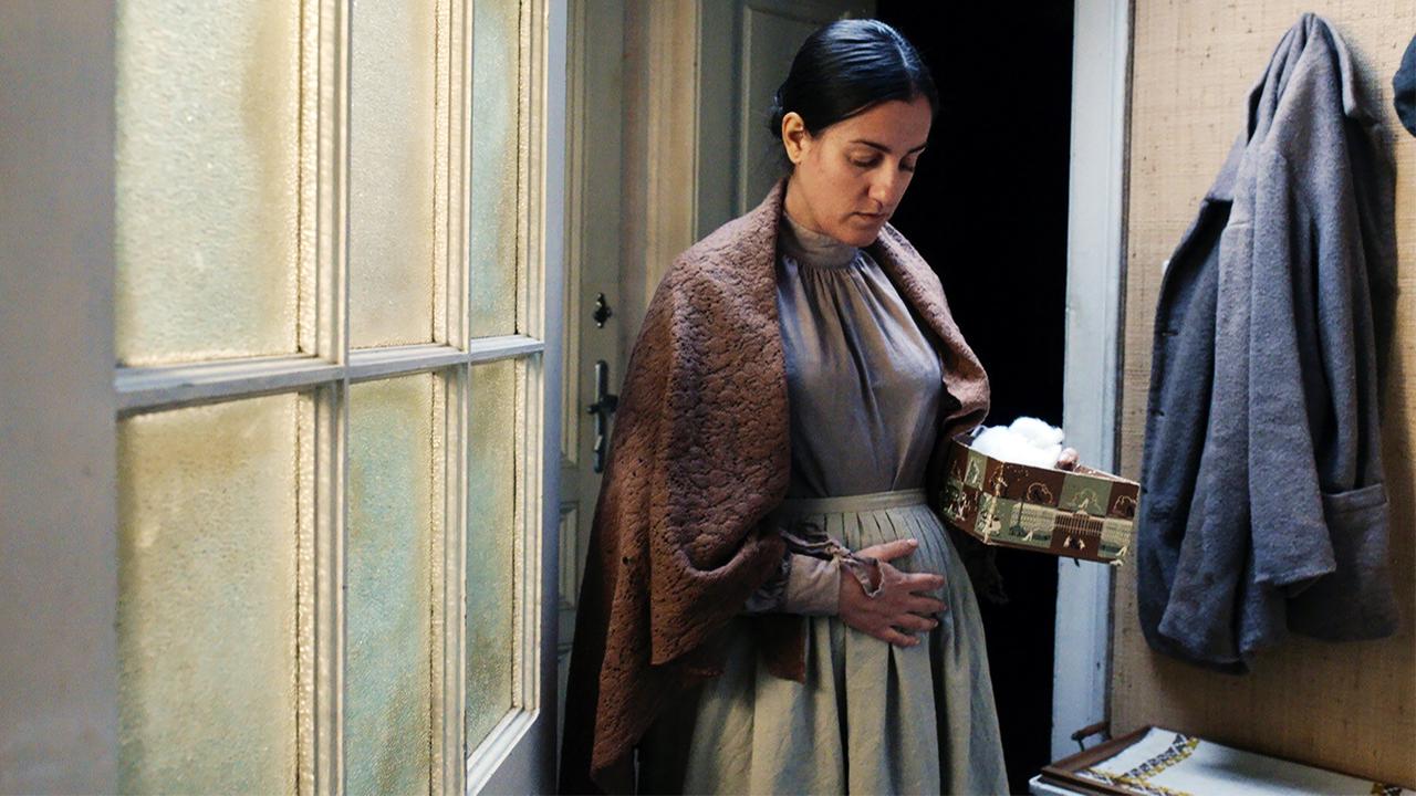 Das Bild zeigt eine Frau, die in einem schlichten Raum steht. Sie trägt ein langes, schlichtes Kleid in gedeckten Farben und hat einen braunen Überwurf um die Schultern gelegt. Ihr Gesicht und ihre Haltung vermitteln eine nachdenkliche oder besorgte Stimmung.   In ihrer linken Hand hält sie eine dekorative Box, die mit Watte gefüllt ist. Der Hintergrund zeigt eine Tür und eine Wand mit einer rauen Textur, an der eine Jacke hängt. Durch ein Fenster auf der linken Seite scheint gedämpftes Licht, das eine sanfte Stimmung erzeugt. Der Raum wirkt einfach und unprätentiös.   Die dargestellte Szene spielt offenbar in einer historischen Umgebung, die möglicherweise die Weihnachtszeit in der Monarchie widerspiegelt.