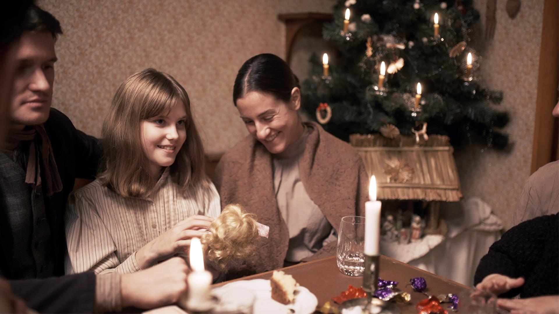 Das Bild zeigt eine weihnachtliche Szene in einem kleinen, bescheidenen Raum. Im Mittelpunkt sitzt eine Familie um einen Tisch, der mit verschiedenen Lebensmitteln, wie Kuchen und Süßigkeiten, gedeckt ist. Auf dem Tisch stehen auch Kerzen, die eine warme, gemütliche Atmosphäre schaffen.  Im Hintergrund sieht man einen Christbaum, der mit kleinen Kerzen und traditionellem Schmuck geschmückt ist. Die Wände des Raumes sind mit einem einfachen, gedämpften Muster dekoriert.   Eine Mutter und eine Tochter, die ein Spielzeug in den Händen hält, strahlen Freude aus. Beide sind in einfache, historische Kleidung gehüllt; die Mutter trägt ein braunes Tuch um die Schultern. Weitere Familienmitglieder sind am Tisch zu sehen, die gut gelaunt wirken und sich miteinander unterhalten. Das Licht der Kerzen taucht die Szene in ein sanftes, warmes Licht und verstärkt die festliche Stimmung.