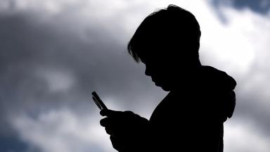 Jugendlicher schaut auf sein Smartphone, Schattenriss vor dunklen Wolken.