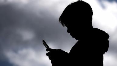 Jugendlicher schaut auf sein Smartphone, Schattenriss vor dunklen Wolken.