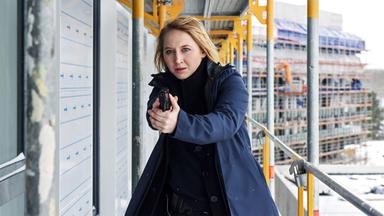 Nora Weiss (Anna Maria Mühe) steht mit gezogener Waffe auf einem Baugerüst und schaut angespannt.  